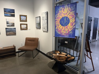 An art gallery interior showcasing various paintings on the wall, featuring seascapes and abstracts, with a colorful sun-themed tapestry hanging. A modern brown chair and a unique wooden table with natural edges are visible in the space.