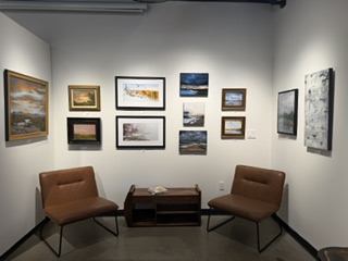 A cozy art gallery corner featuring a wall display of various framed paintings. Two brown chairs are positioned in front of the wall, with a small wooden table between them.