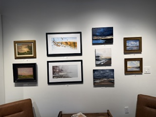 An interior wall displaying a variety of framed artworks, including landscapes and seascapes, arranged in a grid pattern. The artworks feature different colors and styles, with some in traditional frames and others more modern.