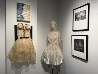 A gallery display featuring two unique dresses on mannequins, one with a structured top and belt, the other with lace detailing, accompanied by two framed photographs on the wall.