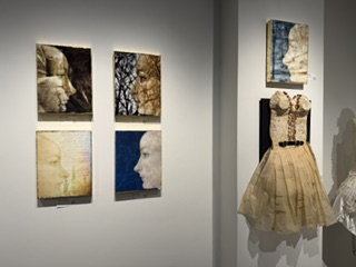 An art gallery display featuring four framed artworks of faces on the left wall and a dress made of paper or fabric displayed on the right wall.