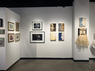 Art gallery interior featuring a variety of framed artworks, including photographs and paintings, alongside a decorative dress displayed on the wall.