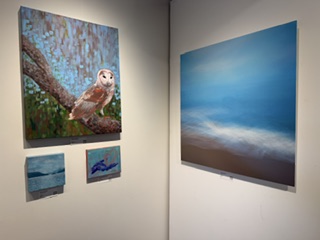 A gallery wall displaying four paintings: a close-up of an owl perched on a branch, a landscape with water, a colorful abstract artwork, and a large blue abstract piece.