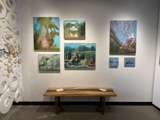 A gallery wall featuring various paintings, including landscapes and an owl, alongside a wooden bench in an art space.