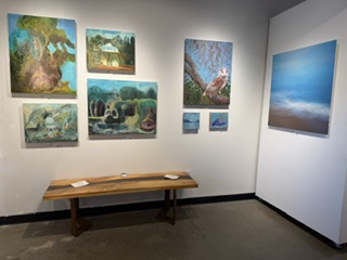 An art gallery featuring several colorful paintings on the wall, including landscapes and abstract works, with a wooden bench in the foreground.