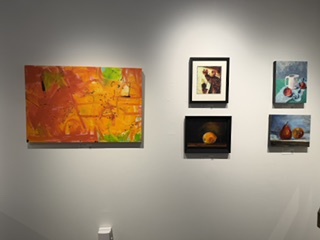 A display of various artworks hanging on a wall, including a large orange abstract painting, smaller framed artworks featuring still life and fruit imagery.