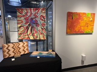 An art gallery display featuring colorful wall art and decorative wooden pieces. On the left, a vibrant textile artwork with a floral design, next to abstract paintings in bright orange and pink hues. Below, two wooden art pieces with a zigzag pattern and a smooth, blue-accented design on a black table.