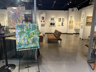 An art gallery with various artworks displayed on the walls, featuring a colorful abstract painting on an easel in the foreground, and a brown sofa in the center of the room.